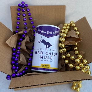 May include: A cardboard box with brown packing paper and purple and gold beaded necklaces. A white can with a black mule illustration and the text "Give Your Food A Kick!"  "EST. 2023" "MAD CAJUN MULE" "SEASONING" "Net Weight 7 oz. (198g)" is in the center of the box.