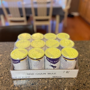 May include: A 12-pack of Mad Cajun Mule seasoning in yellow and purple containers with a yellow lid. The containers are arranged in a cardboard box with the text "Mad Cajun Mule" and "7 oz" printed on the front. The box also has a "Best By 02/02/27" date printed on the bottom.