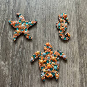 Puede incluir: Tres adornos de madera decorados con cuentas y lentejuelas de colores. Los adornos tienen forma de estrella de mar, caballito de mar y tortuga.