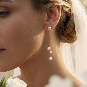 Op de afbeelding: Elegante gouden oorbellen met drie bungelende parels. De oorbellen zijn bevestigd aan een kleine parelknop. De oorbellen worden gedragen door een persoon met blond haar en een witte sluier. De achtergrond is vervaagd met witte bloemen.