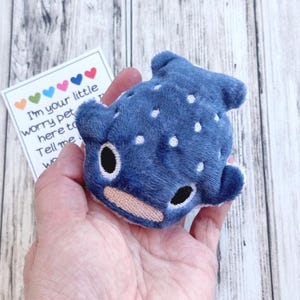May include: A blue whale shark plush toy with white spots and black eyes. The toy is held in a hand, with a small card that reads "I'm your little worry pet..." with colourful hearts.