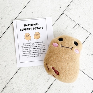 Puede incluir: Una patata de peluche marrón con una cara sonriente y mejillas rosadas. La patata sostiene una tarjeta que dice "Patata de apoyo emocional" e incluye un mensaje sobre estar ahí para alguien cuando necesita un hombro en el que apoyarse.