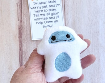 May include: A small, white plush toy with a blue belly and black eyes is held in a hand. A card with the text "I'm your little worry pet and I'm here to stay. Tell me all your worries and I'll help them go away!" is in the background.