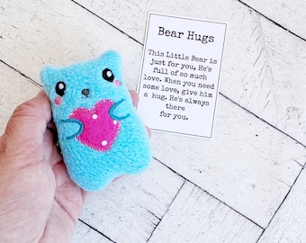May include: A small, light blue plush bear with a pink heart and black eyes. The bear is held in a hand, with a card that reads "Bear Hugs" and a message about love and hugs.