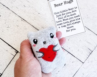 May include: A small gray plush bear with a red heart on its chest. The bear is holding the heart with its paws. The bear is sitting in a hand. A card with the text "Bear Hugs" is in the background. The card says "This Little Bear is just for you. He's full of so much love. When you need some love, give him a hug. He's always there for you."