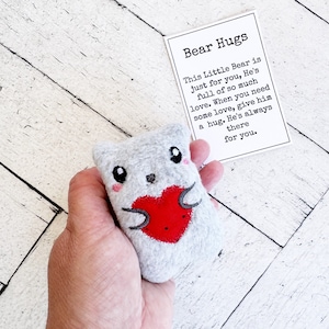 Puede incluir: Un pequeño oso de peluche gris con un corazón rojo en el pecho. El oso está sosteniendo el corazón con sus patas. El oso está sentado en una mano. Una tarjeta con el texto "Bear Hugs" está en el fondo. La tarjeta dice "This Little Bear is just for you. He's full of so much love. When you need some love, give him a hug. He's always there for you."