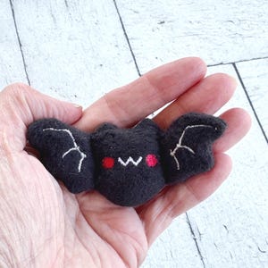 Pode incluir: Um pequeno morcego de pelúcia preto com detalhes bordados, incluindo contornos de asas brancos, uma boca branca em forma de w e duas bochechas vermelhas circulares. Esta criatura macia e felpuda tem um corpo arredondado e asas estendidas.