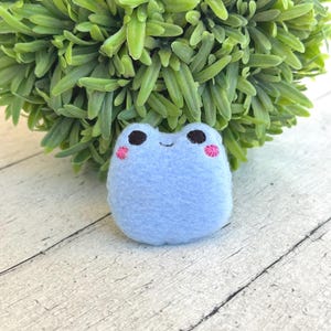 May include: A light blue felt frog with black eyes and pink cheeks. The frog is sitting on a white wooden surface with a green plant in the background. The frog is a handmade item.