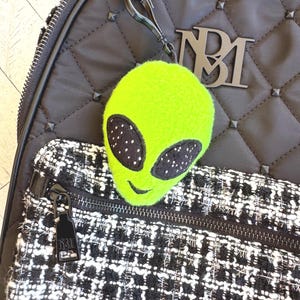 May include: A bright neon green plush alien head keychain with large black oval eyes featuring white speckles and a small stitched smile. The keychain is attached to a black quilted backpack with a black and white tweed front pocket and NBM logo.