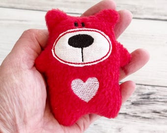May include: A small red plush bear with a white heart and a white face with a black nose and eyes.