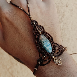 Puede incluir: Una pulsera de mano de macramé marrón con una piedra central ovalada de color azul y gris. El diseño incluye intrincados patrones de cordón anudado, pequeñas cuentas metálicas y un pequeño dije metálico colgante en la zona de la muñeca.