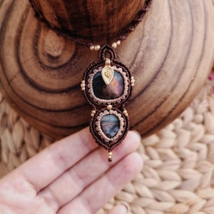 Puede incluir: Un collar de macramé hecho a mano con dos colgantes de piedras preciosas oscuras e iridiscentes. Los colgantes están enmarcados con intrincados nudos de macramé en tonos marrones y beige, acentuados con cuentas doradas. El collar se exhibe sobre una superficie de madera.