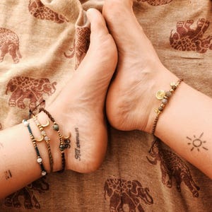Può includere: Braccialetti alla caviglia con ciondoli a forma di luna e perline adornano le caviglie. I piedi poggiano su un tessuto marrone fantasia con motivi di elefanti. Un tatuaggio a forma di sole è visibile su una caviglia e un tatuaggio con scritta sull'altra.
