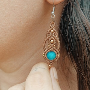 Peut inclure: Une boucle d'oreille en macramé composée d'un cordon marron en forme de goutte avec des nœuds complexes, de petites perles dorées métalliques et une pierre ronde de couleur turquoise à la base, suspendue à un crochet en métal argenté.