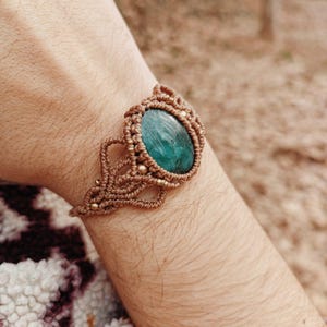 Puede incluir: Pulsera de macramé hecha a mano con una piedra ovalada de color turquesa. La pulsera está elaborada con cordón marrón y pequeñas cuentas doradas, mostrando intrincados patrones de nudos. La pulsera se lleva en la muñeca.