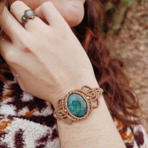 May include: A macrame bracelet with a large, oval, emerald-green stone at the center. The bracelet is made of tan-colored cord and features intricate knotting details. A matching ring with a smaller stone is also visible.