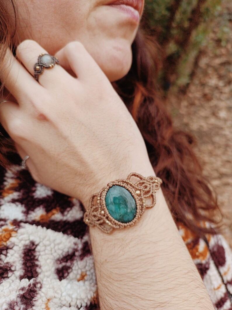 May include: A macrame bracelet with a large, oval, emerald-green stone at the center. The bracelet is made of tan-colored cord and features intricate knotting details. A matching ring with a smaller stone is also visible.