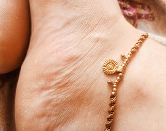 May include: A gold-coloured anklet featuring a sun-shaped charm and decorative beads. The anklet is worn on an ankle, highlighting the detailed design and craftsmanship. The anklet is a piece of jewellery.