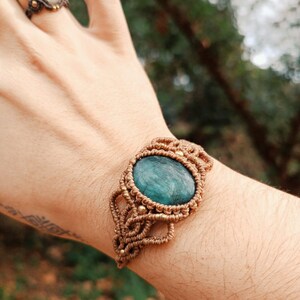 May include: A macrame bracelet with a large, oval, teal-colored stone at its center. The bracelet is made of tan-colored cord and features intricate knotting details. A matching ring with a light-colored stone is also visible.