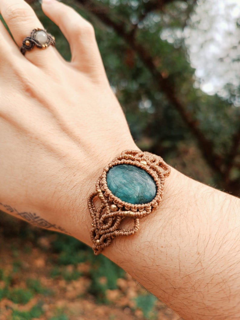 May include: A macrame bracelet with a large, oval, teal-colored stone at its center. The bracelet is made of tan-colored cord and features intricate knotting details. A matching ring with a light-colored stone is also visible.