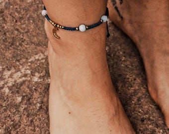 Può includere: Braccialetto alla caviglia con cordino blu scuro, perline color oro e un ciondolo a forma di mezzaluna. Sono presenti anche perline azzurre. Il gioiello è indossato sulla caviglia.