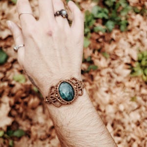 May include: A macrame bracelet with a large, oval, teal-colored stone at the center. The bracelet is made of a light brown cord and is worn on a wrist. Several rings are visible on the hand.