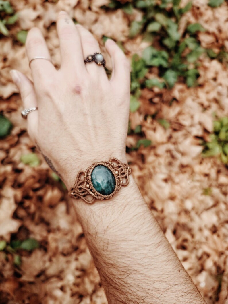 May include: A macrame bracelet with a large, oval, teal-colored stone at the center. The bracelet is made of a light brown cord and is worn on a wrist. Several rings are visible on the hand.