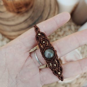 Puede incluir: Pulsera de macramé marrón hecha a mano, con una piedra gris redonda en el centro. Adornada con pequeñas cuentas doradas. Los intrincados nudos y los colores neutros le dan una estética bohemia. Una pieza de joyería única.