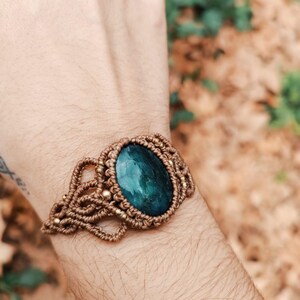 May include: A handmade macrame bracelet featuring a large, oval, dark teal gemstone. The bracelet is crafted with tan-colored macrame cord and accented with small gold-colored beads. The gemstone is approximately 1.5 inches long.