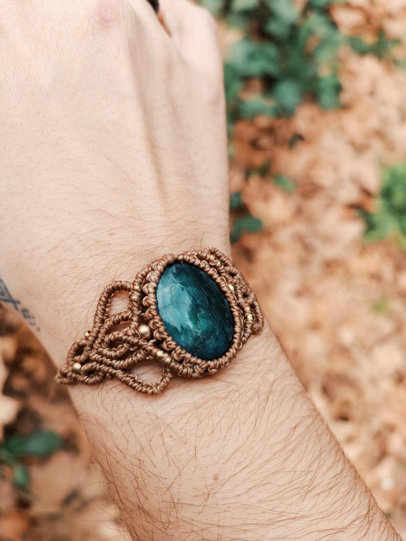 May include: A handmade macrame bracelet featuring a large, oval, dark teal gemstone. The bracelet is crafted with tan-colored macrame cord and accented with small gold-colored beads. The gemstone is approximately 1.5 inches long.