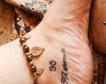 May include: Ankle bracelet featuring brown beads and gold-toned leaf charms. The bracelet adorns an ankle with a tattoo reading "www.mypeace.com" and the number "98". The background is a patterned fabric.
