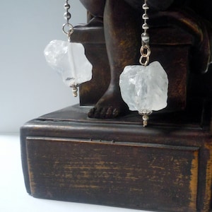 May include: Two clear quartz crystal pendants hang from silver chains. The raw, irregular quartz stones are suspended from decorative silver findings. The pendants are displayed against a dark brown, wooden background, with a statue's foot visible.