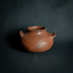 Puede incluir: Una olla de cerámica hecha a mano, de color marrón rojizo, con dos pequeñas asas. La olla tiene un cuerpo redondeado y una abertura ancha. La superficie muestra ligeras imperfecciones, lo que sugiere una calidad rústica y artesanal. La olla está sobre un fondo oscuro.