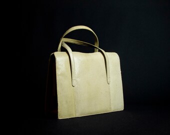 May include: A light beige leather handbag with two top handles. The bag has a textured surface and a slightly rounded shape.