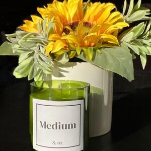 Pu&ograve; includere: Un barattolo di vetro verde per candele con un'etichetta bianca che recita "Medium 8 oz" e uno stoppino bianco. La candela &egrave; appoggiata su una superficie nera davanti a un mazzo di girasoli gialli e foglie verdi.