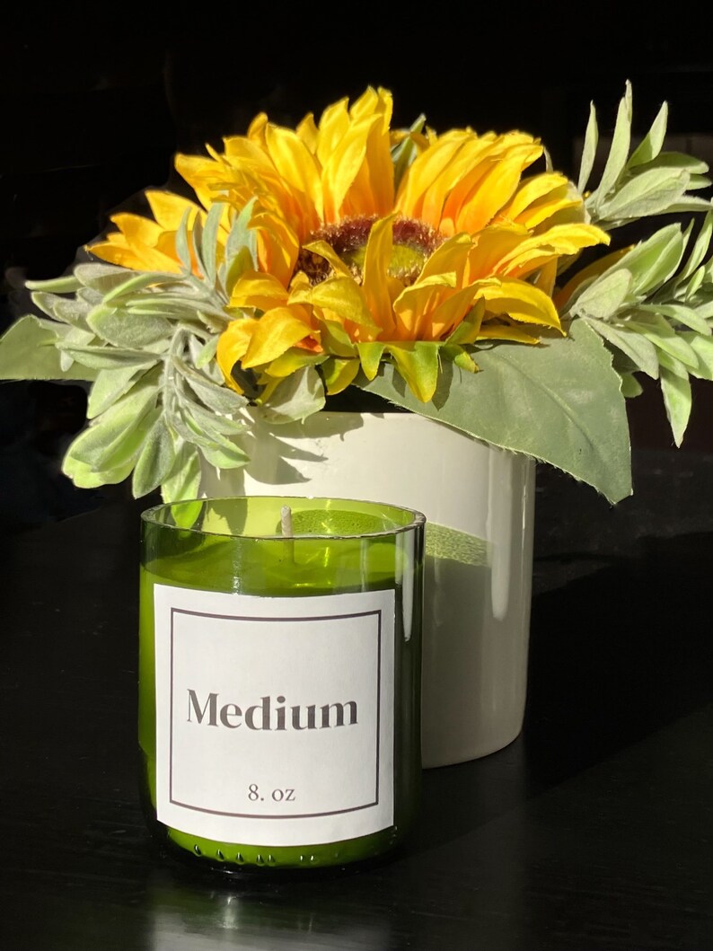 Pu&ograve; includere: Un barattolo di vetro verde per candele con un'etichetta bianca che recita "Medium 8 oz" e uno stoppino bianco. La candela &egrave; appoggiata su una superficie nera davanti a un mazzo di girasoli gialli e foglie verdi.