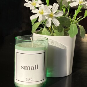 Pu&ograve; includere: Un piccolo barattolo di vetro verde per candele con un'etichetta bianca che dice "small" e "5.5 oz". La candela &egrave; accesa e la cera &egrave; sciolta. La candela &egrave; su una superficie nera accanto a un vaso bianco con fiori bianchi.
