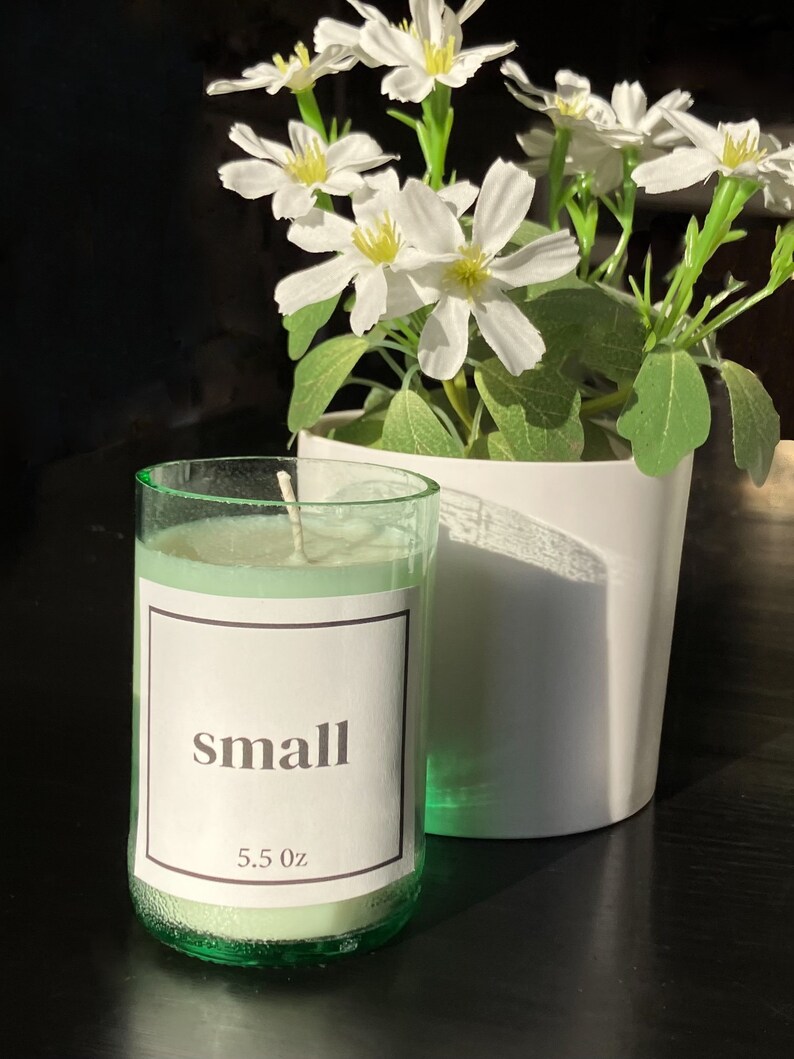 Pu&ograve; includere: Un piccolo barattolo di vetro verde per candele con un'etichetta bianca che dice "small" e "5.5 oz". La candela &egrave; accesa e la cera &egrave; sciolta. La candela &egrave; su una superficie nera accanto a un vaso bianco con fiori bianchi.