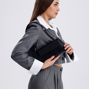 Puede incluir: Una mujer con un blazer gris y una camisa blanca sostiene un bolso de hombro negro.