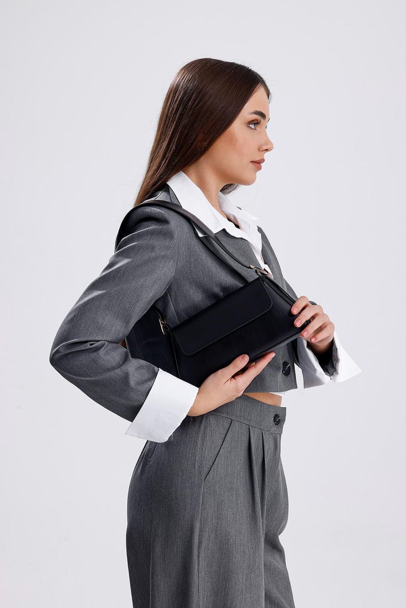 Puede incluir: Una mujer con un blazer gris y una camisa blanca sostiene un bolso de hombro negro.