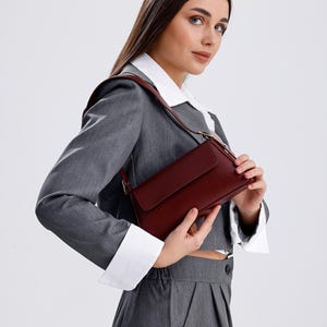 May include: A woman wearing a grey suit with a white collared shirt and a burgundy shoulder bag.