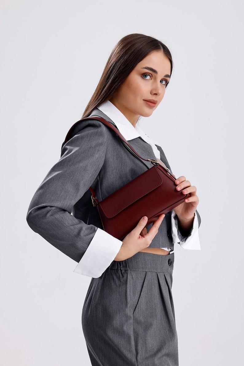 May include: A woman wearing a grey suit with a white collared shirt and a burgundy shoulder bag.