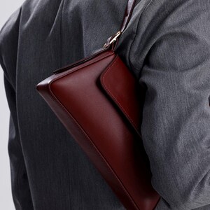May include: A burgundy leather shoulder bag with a flap closure and a gold-toned buckle. The bag is worn over the shoulder of a person wearing a grey blazer.
