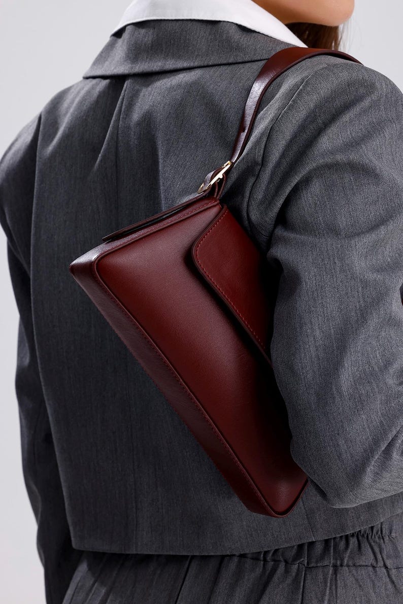 May include: A burgundy leather shoulder bag with a flap closure and a gold-toned buckle. The bag is worn over the shoulder of a person wearing a grey blazer.