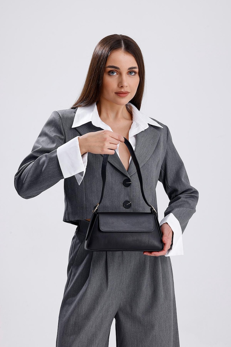May include: A woman wearing a gray blazer and pants with a white collared shirt underneath. She is holding a black handbag with a gold clasp.