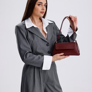May include: A woman wearing a grey suit jacket and trousers with a white collared shirt underneath. She is holding a burgundy leather handbag.
