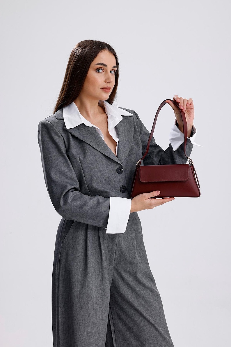 May include: A woman wearing a grey suit jacket and trousers with a white collared shirt underneath. She is holding a burgundy leather handbag.