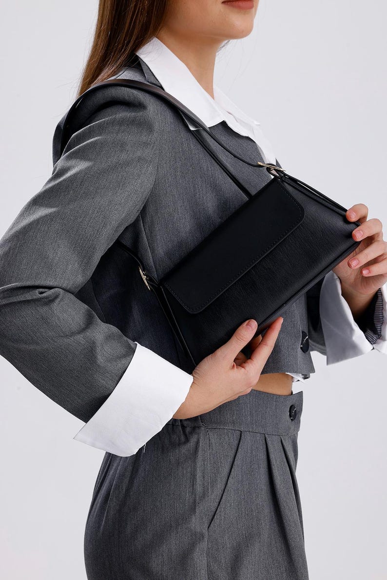 Puede incluir: Un bolso de hombro de cuero negro con cierre de solapa. El bolso tiene una hebilla dorada y una correa larga.