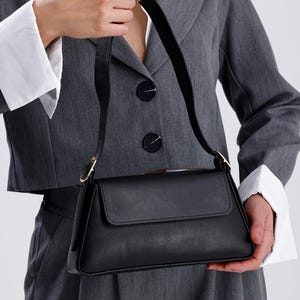 Puede incluir: Un bolso de hombro de cuero negro con cierre de solapa y hebilla dorada. El bolso tiene una forma estructurada y una correa corta.
