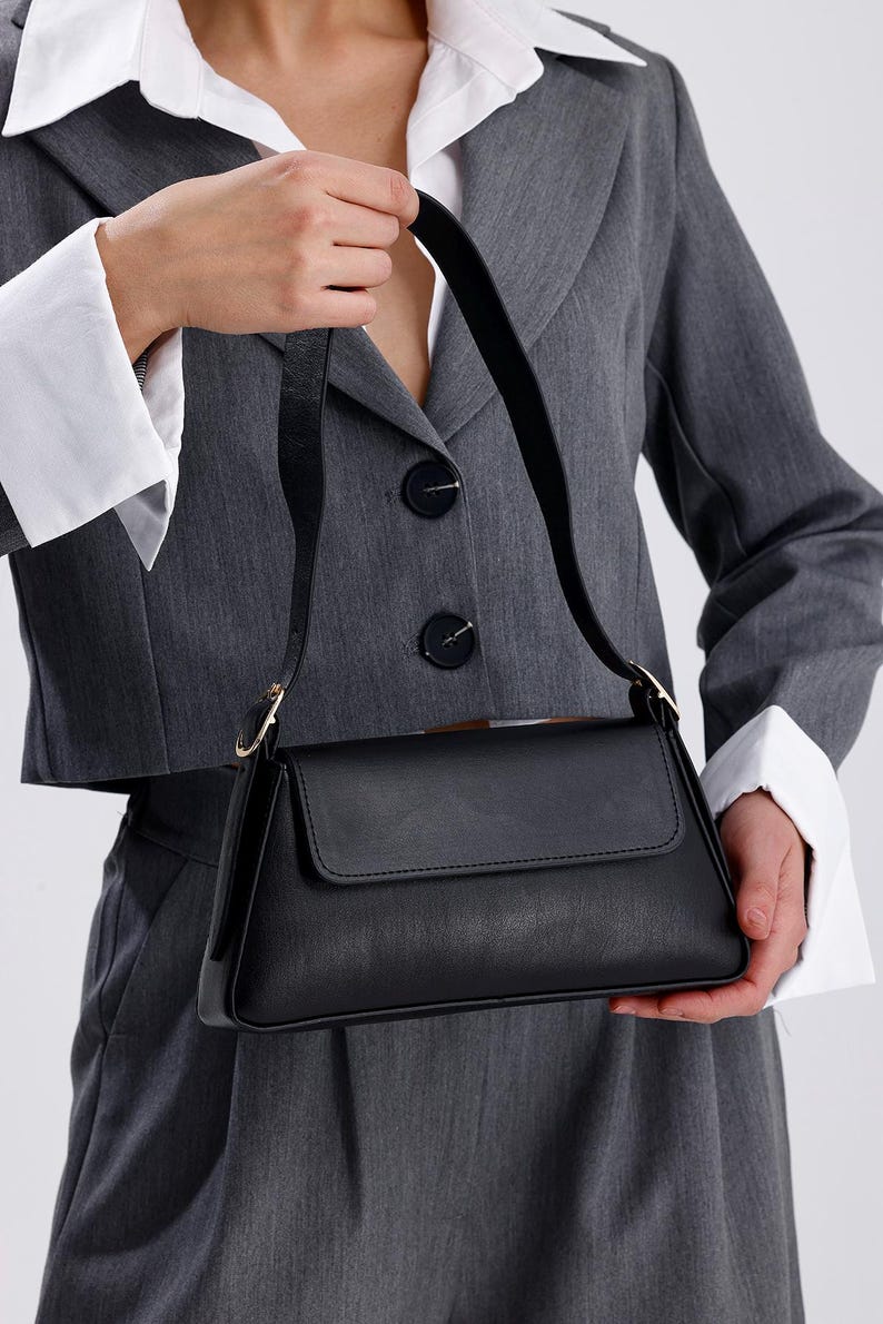 Puede incluir: Un bolso de hombro de cuero negro con cierre de solapa y hebilla dorada. El bolso tiene una forma estructurada y una correa corta.